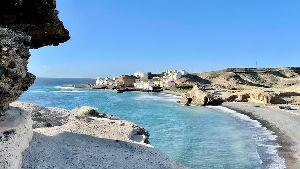 A coastal town with buildings along the shoreline, a rocky foreground, and clear blue waters.