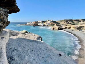 Une ville côtière avec des bâtiments qui bordent le littoral, un premier plan rocheux et des eaux d’un bleu limpide.