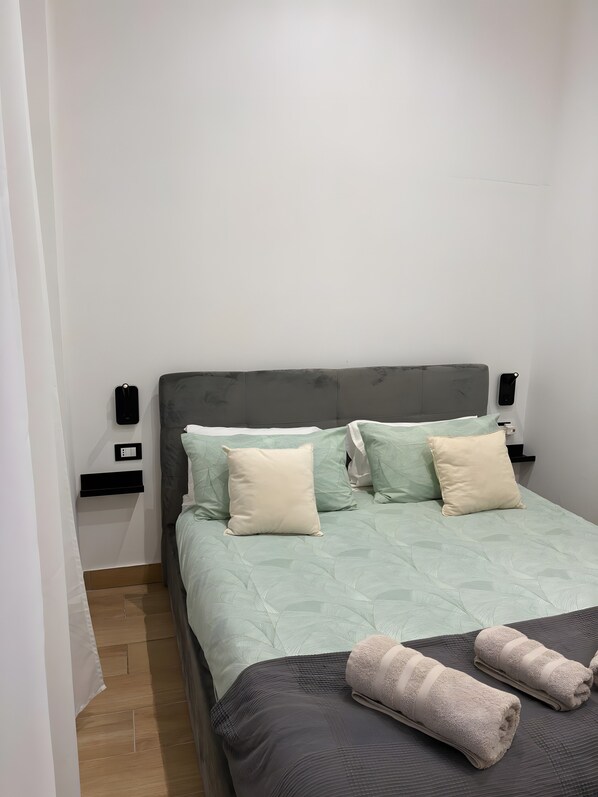 Un lit bien fait, avec une tête de lit grise, deux oreillers, une table de chevet sur laquelle se trouve un téléphone et une petite étagère, et un plancher en bois.