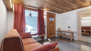 A cozy room with a wooden ceiling, a sofa with a plaid pillow, a wooden door with a window, and a small table with a lamp.