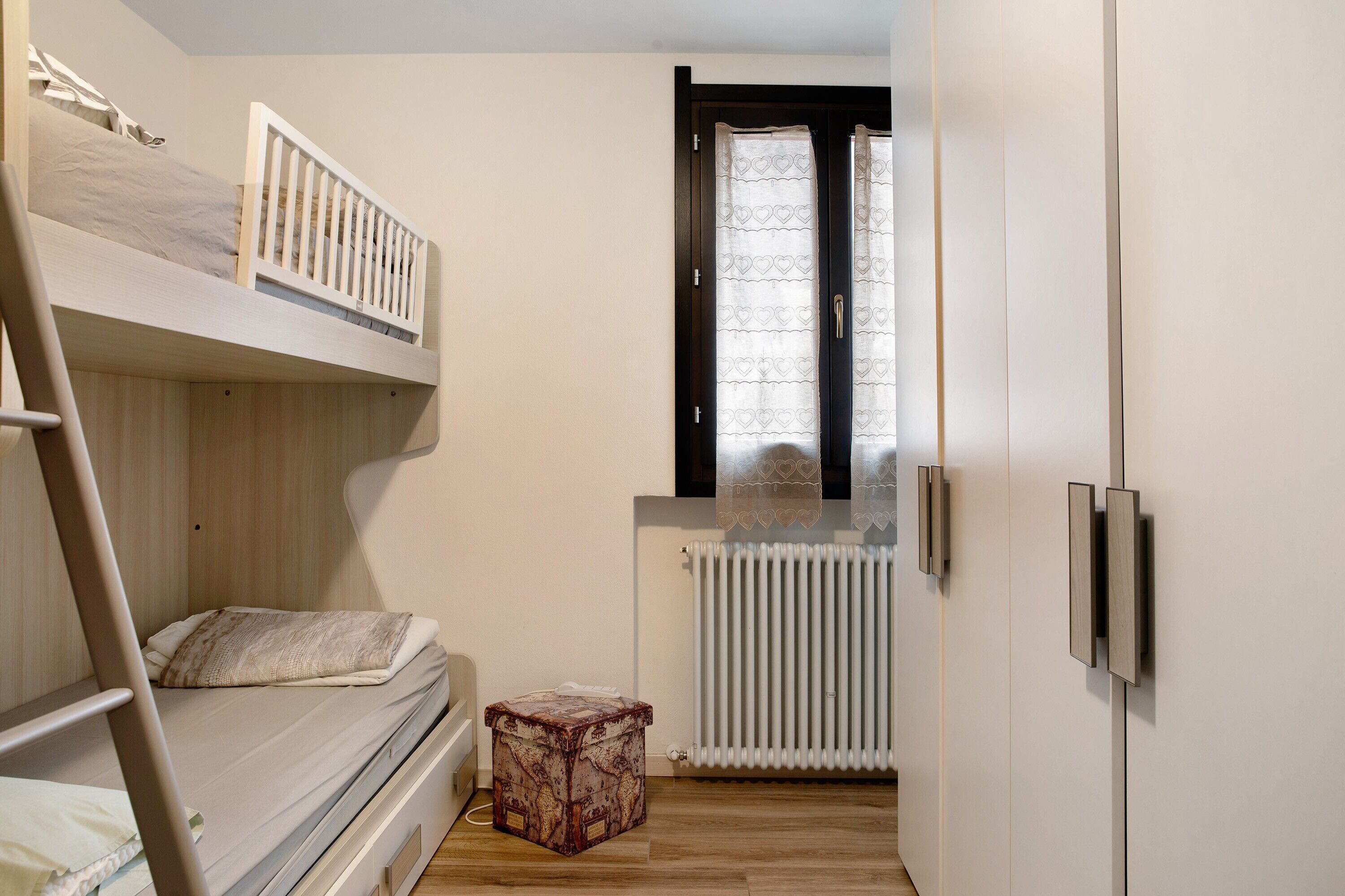 A bunk bed room with a ladder, a radiator, a small wooden stool, and a window with curtains.