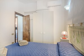Un dormitorio con una cama azul acolchada, una puerta de madera, un armario blanco, una mesita de noche con lámpara y un aire acondicionado instalado en la pared.
