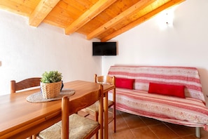 Una mesa de comedor de madera con sillas, una cesta tejida con una planta, un sofá con cojines rojos y un televisor montado en la pared.