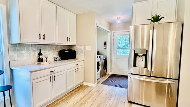 A modern kitchen with white cabinets, a stainless steel refrigerator, a coffee maker, and a washing machine.
