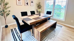 A dining area with a wooden table, black chairs, and a vase with flowers. There are framed artworks on the wall and a window with a view of greenery.