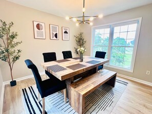 A dining area with a wooden table, black chairs, and a vase with flowers. There are framed artworks on the wall and a window with a view of greenery.