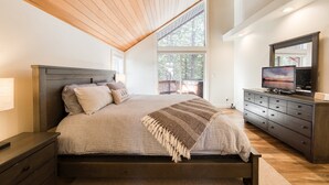 A bedroom with a large bed, a dresser, a television, and a window with a view of trees.