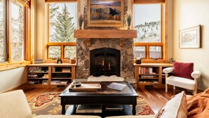 Una sala de estar acogedora con chimenea de piedra, estanterías de madera, una mesa de centro y vistas a un paisaje nevado a través de amplias ventanas.