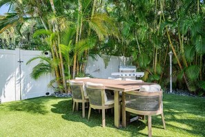 A backyard with a wooden table and chairs, a grill, and lush greenery.