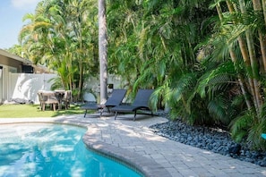A swimming pool with lounge chairs, surrounded by palm trees and a patio area. - Cozy Cabana West – AMI Pet Friendly Bungalow w/ Heated Pool & Walk to Beach (Holmes Beach)