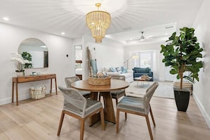 A modern living room with a round wooden dining table, wicker chairs, a large mirror, and a chandelier. - Cozy Cabana West – AMI Pet Friendly Bungalow w/ Heated Pool & Walk to Beach (Holmes Beach)