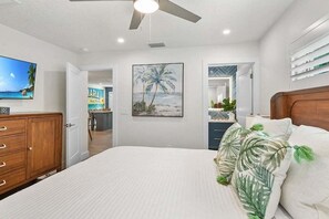 A bedroom with a bed, a ceiling fan, a TV, a painting, and a view of the kitchen. - Cozy Cabana West – AMI Pet Friendly Bungalow w/ Heated Pool & Walk to Beach (Holmes Beach)