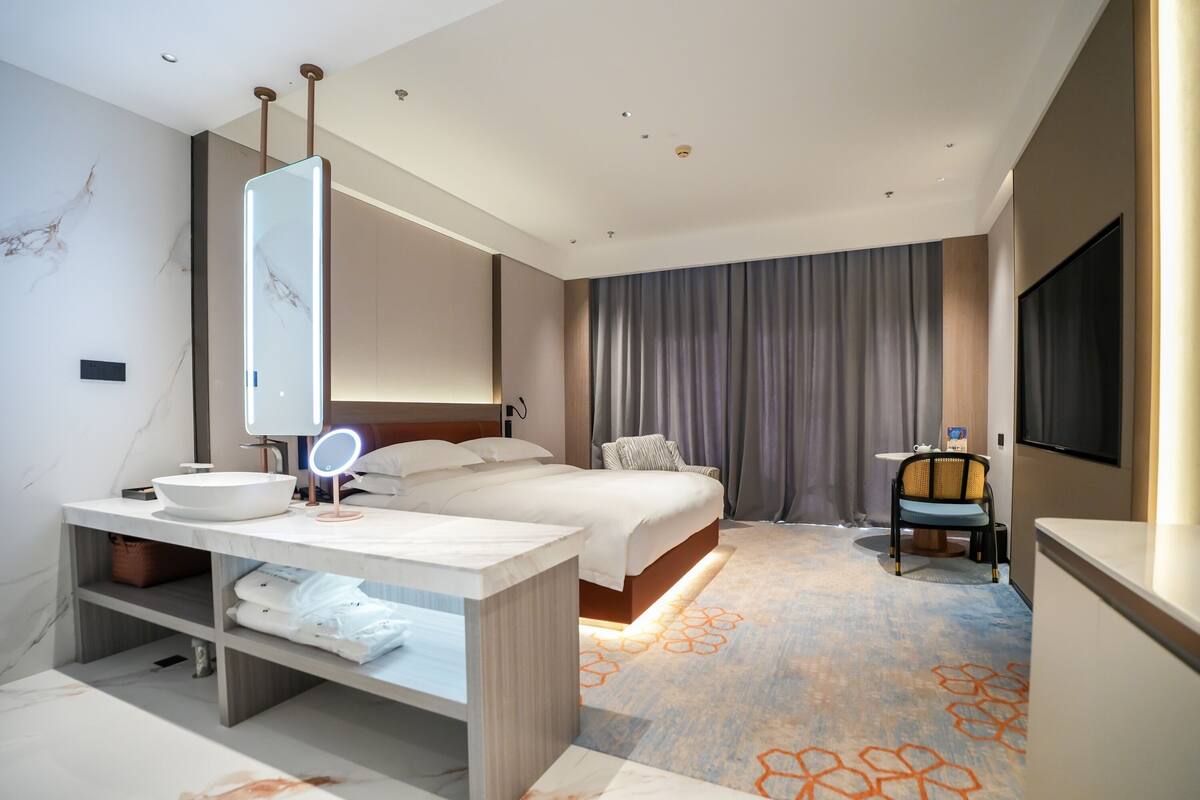 A modern hotel room with a large bed, a bedside table with a mirror, a flat-screen TV, and a bathroom with a marble wall and a sink.