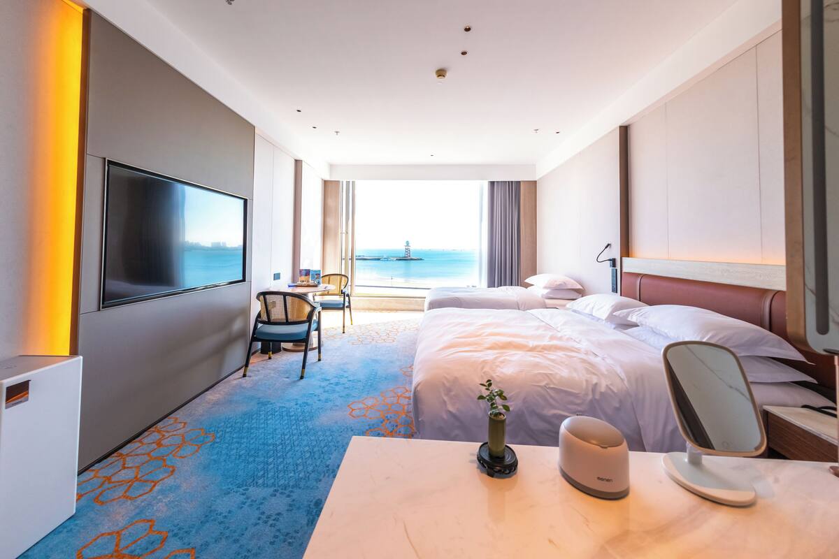 A modern hotel room with a large bed, a TV, a desk with a chair, and a view of the sea.