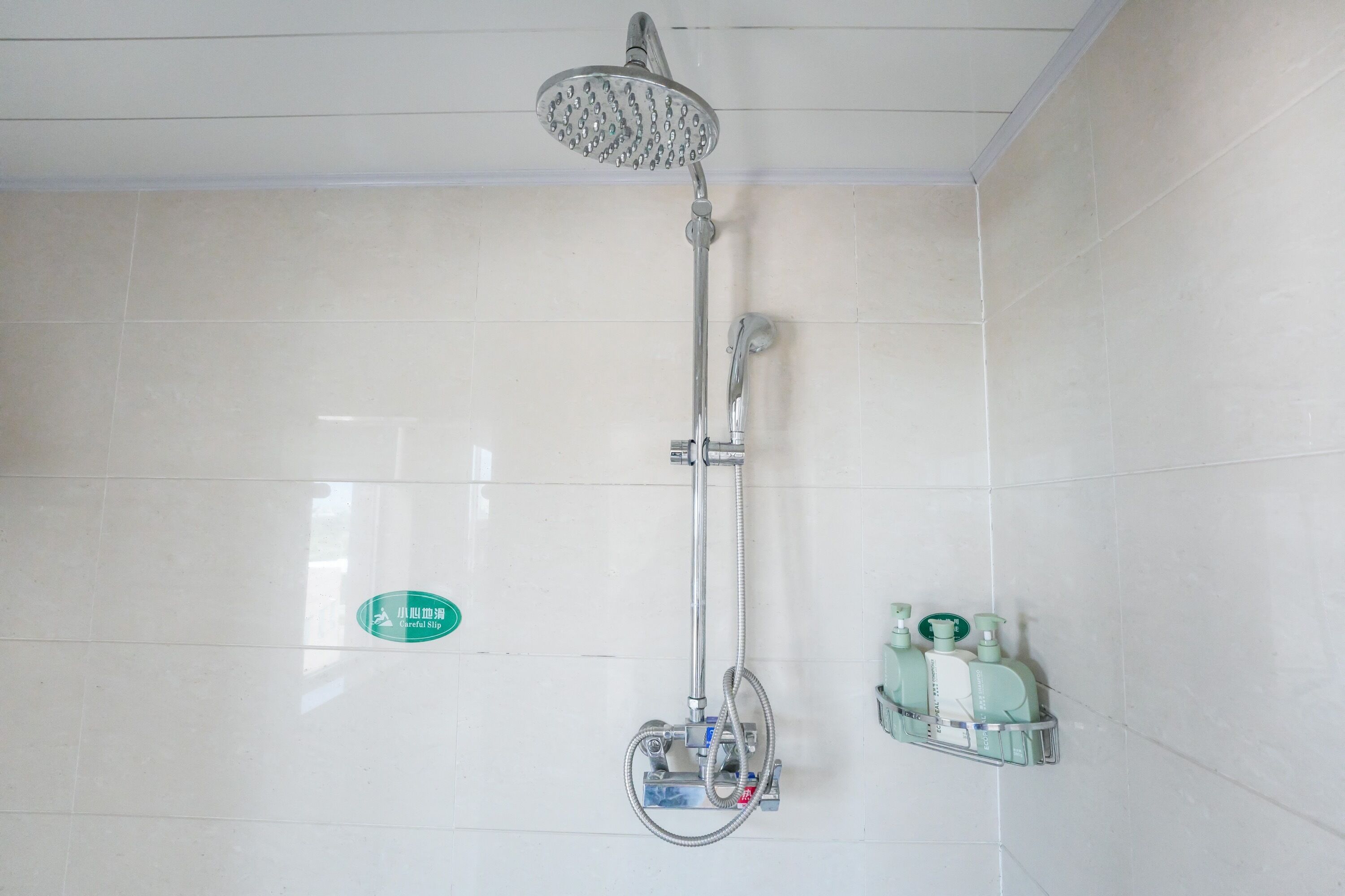 Een douche met een regendouche, een handdouche en een muurkranen. Er hangt een groen bord aan de muur en een zeepdispenser rechts op de muur.