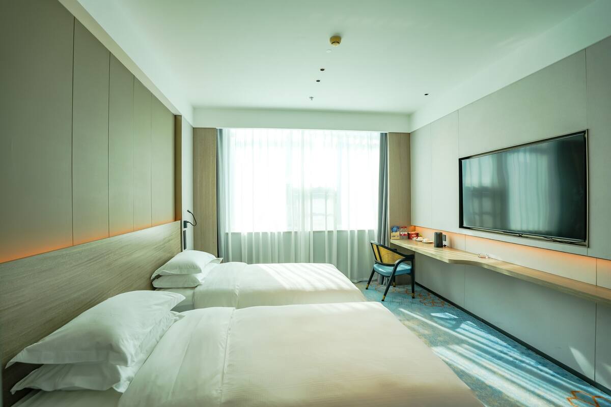 A hotel room with two beds, a desk, a chair, a flat-screen TV, and a large window with curtains.