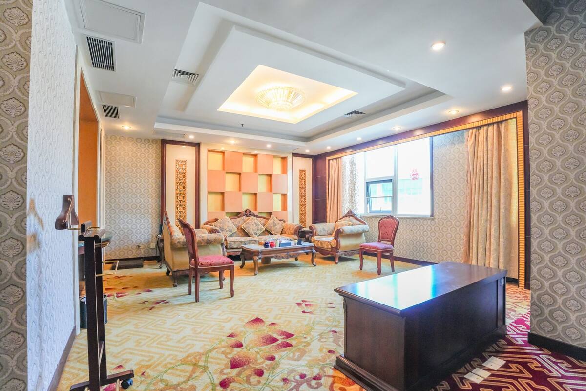 A hotel room with a large sofa set, a wooden coffee table, and a desk. The room has patterned wallpaper and a carpet with a floral design.