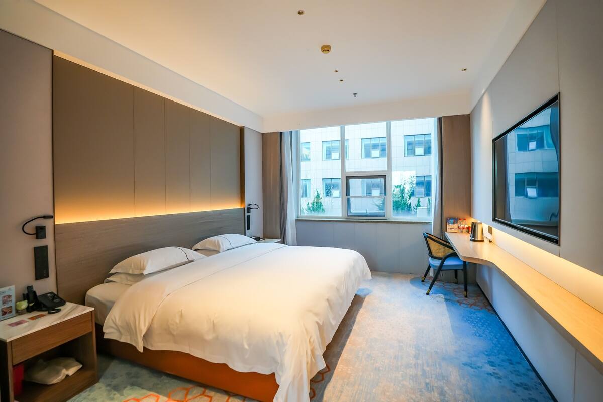 A modern hotel room with a large bed, a desk, and a view of the building outside.