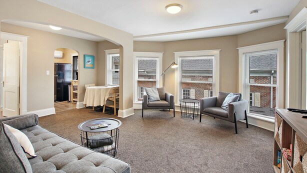 A spacious living room with a grey sofa, two armchairs, a coffee table, and a dining area with a table and chairs. - Neighboring suites in central heart of town! Perfect for couples! Walk to Town, Balcony, & Parking (Lake Geneva)
