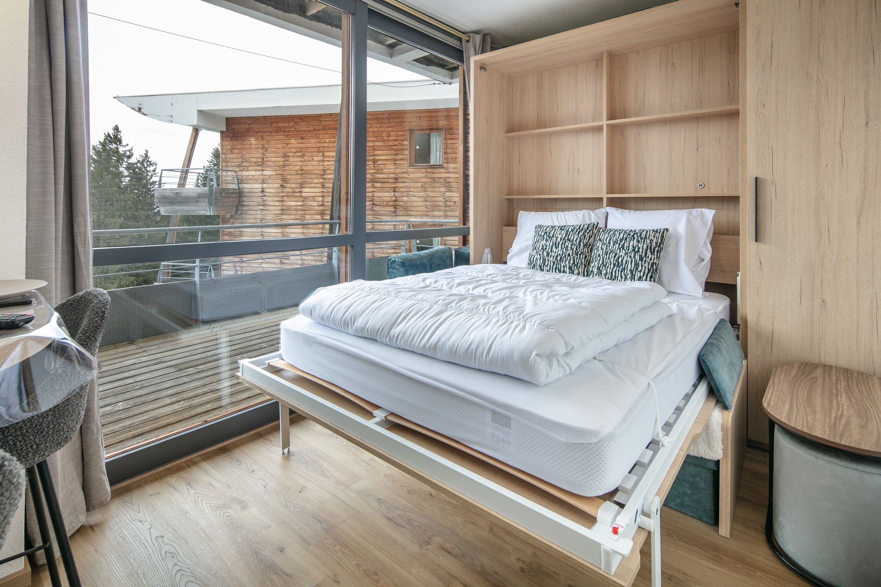 A modern bedroom with a bed, built-in wooden wardrobe, and a balcony with a view of a wooden cabin.