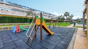 A playground with a slide and swings, surrounded by a paved area and a building with a blue roof in the background.