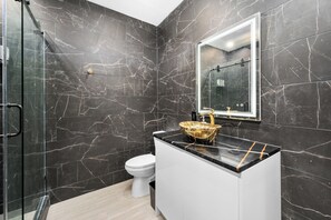 Un baño moderno con paredes de mármol oscuro, cabina de ducha de vidrio, inodoro blanco y lavamanos con detalles dorados.