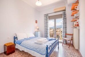Une chambre à coucher comprenant un lit, une chaise, une table de chevet, une fenêtre avec des rideaux et une vue sur des immeubles.