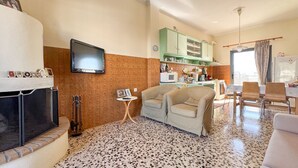 A living room with a fireplace, a television, a dining area with chairs, and a kitchen in the background.