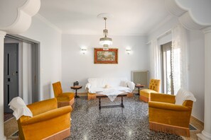 Une pièce comprenant un lit, deux fauteuils, une table d’appoint, un lustre et un tableau accroché au mur.