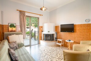 Ein Wohnzimmer mit Sofa, Sesseln, Kamin, Fernseher und Blick durch eine Glastür.