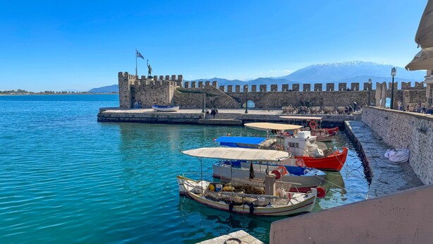 A harbor with boats docked, a stone wall, and a flag on a tower.