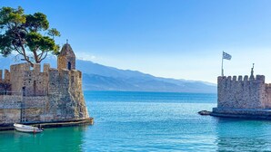 A historic stone fortress with a flag, a clear blue sea, and mountains in the background.