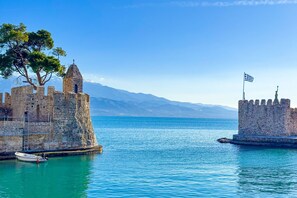 A historic stone fortress with a flag, a clear blue sea, and mountains in the background.