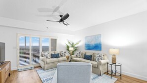 A living room with a beige sectional sofa, a grey armchair, a ceiling fan, a TV on a wooden cabinet, and a view through the glass doors.