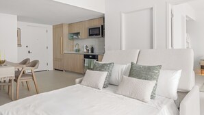 Un dormitorio moderno y minimalista con una cama grande, una pequeña zona de comedor y una kitchenette al fondo.