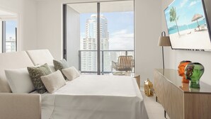 Una habitación de hotel moderna con una cama grande, un televisor que muestra una escena de playa, un balcón con vistas a la ciudad y una pequeña mesa con una lámpara y objetos decorativos.