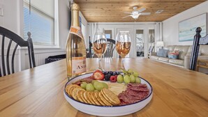 A dining table with a bottle of champagne, two glasses of wine, and a charcuterie board with fruits and crackers. - Steps to Sand on Private Beach | Cozy 2BR Cottage (Gulf Shores)