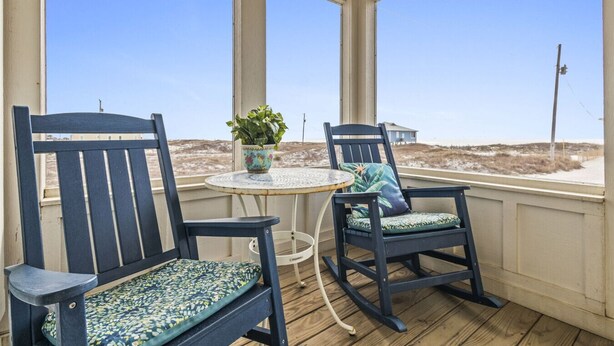 A covered outdoor seating area with two rocking chairs and a table, overlooking a beach. - Steps to Sand on Private Beach | Cozy 2BR Cottage (Gulf Shores)