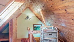 Un interior de madera con techo inclinado, una lámpara fijada a la pared y una silla de mimbre.