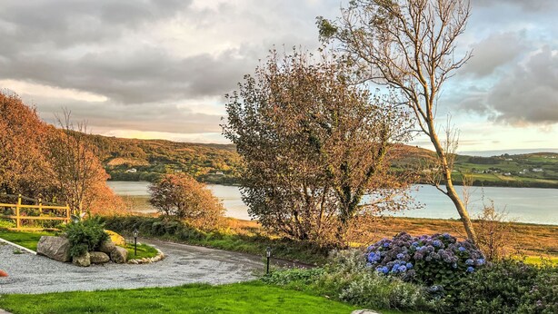 Una vista panorámica de un lago con un sendero de piedra que conduce a una zona cubierta de césped, una valla de madera y un arbusto de flores azules.