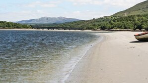 Una playa con un bote en la arena, un puente al fondo y colinas con árboles.