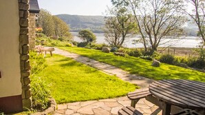 Un patio de piedra con una mesa de madera y bancos que da a una zona verde con un lago y árboles.