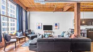 A modern living room with a sectional sofa, a coffee table, and a TV. There is a kitchen area with a microwave and a stove. The room has large windows and a brick wall. - Stylish Industrial Loft in Downtown St. Louis (St. Louis)