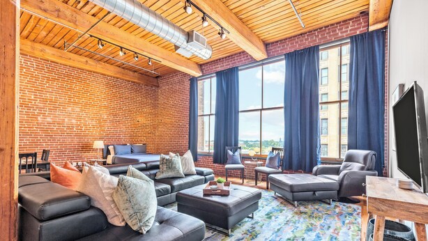 A spacious living area with a brick wall, large windows with curtains, a black leather sofa set, a coffee table, and a television on a wooden stand. - Stylish Industrial Loft in Downtown St. Louis (St. Louis)