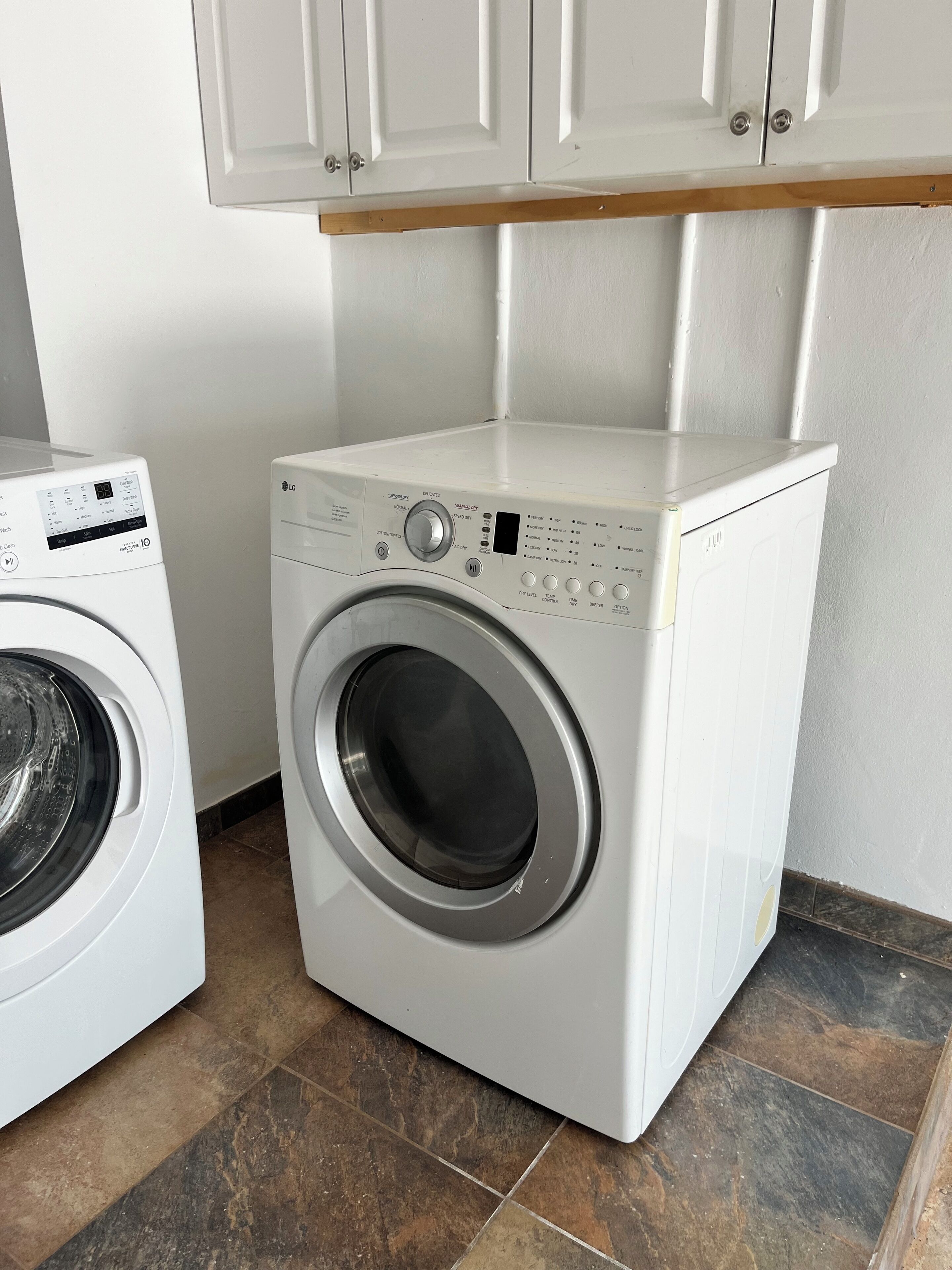 A compact, white front-loading washing machine with a digital display and control panel, situated in a small laundry room with tiled flooring and white cabinetry above.