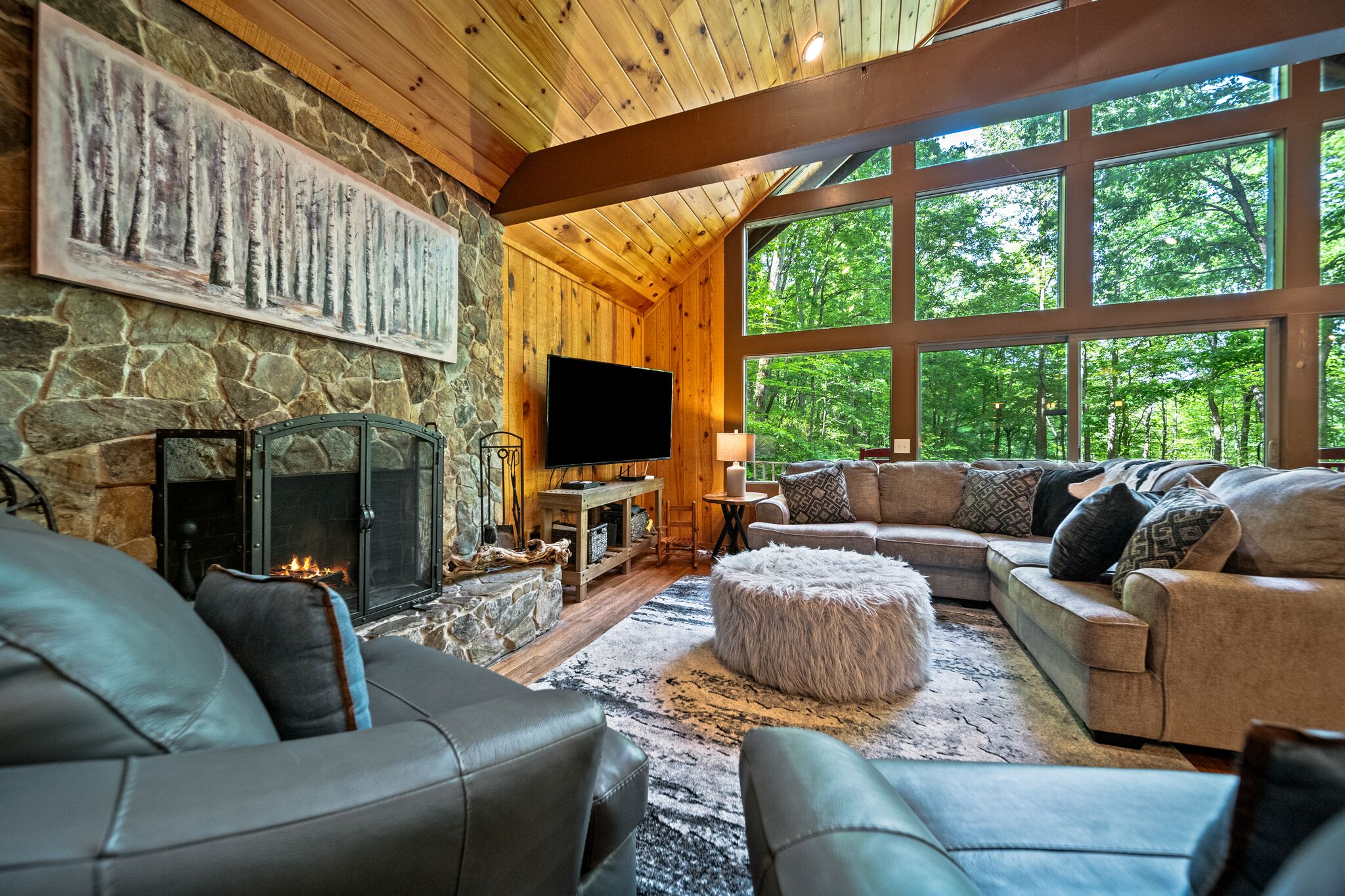 A cozy living room with a stone fireplace, a large TV, and a view of the forest through large windows.