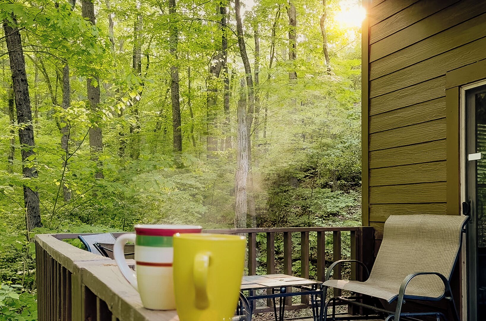 A deck with a view of a forest, a table with chairs, and a yellow mug.