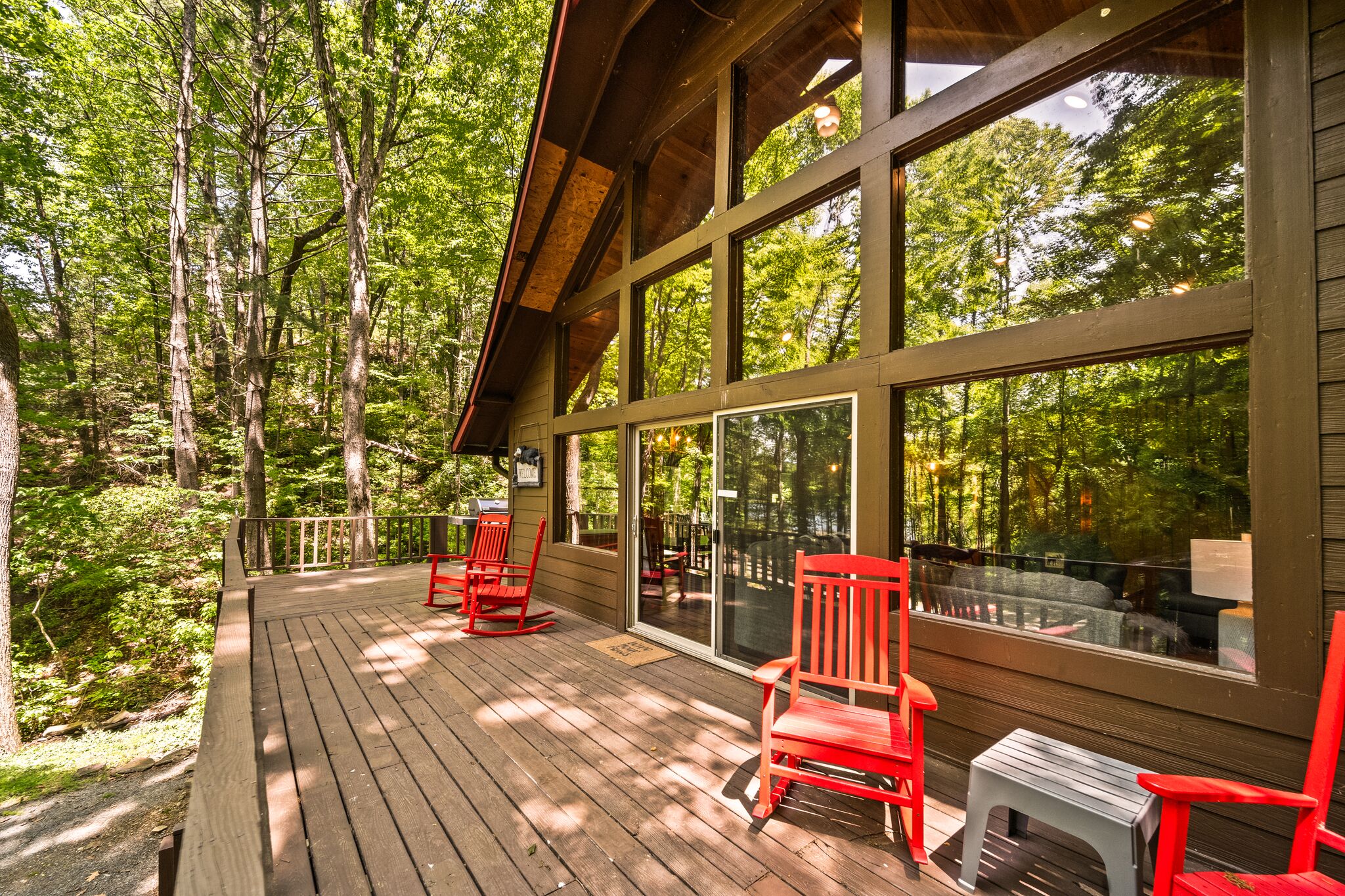 A wooden deck with red chairs and a small table, overlooking a forested area.