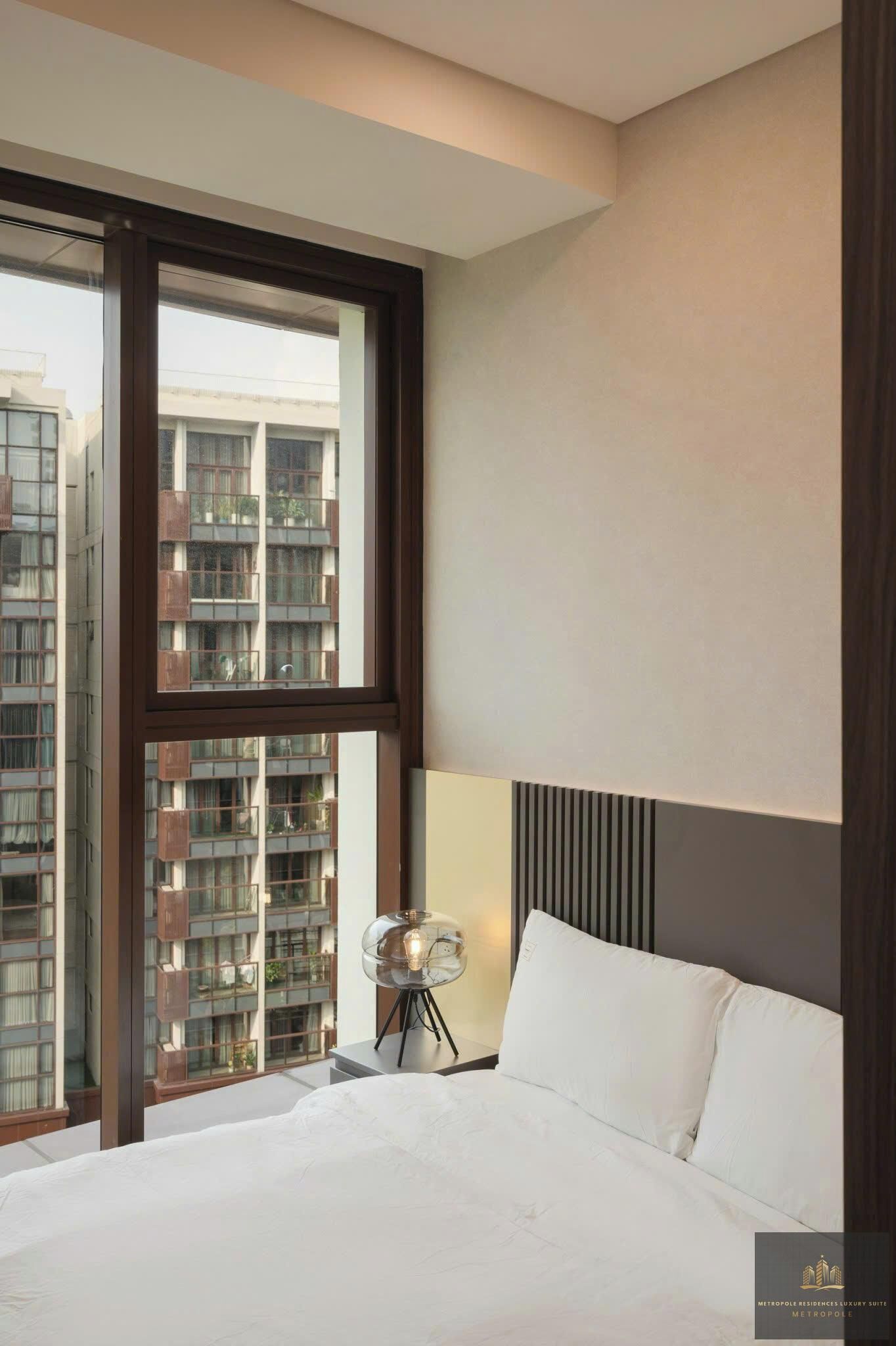 A hotel room with a large window, a bed, a bedside lamp, and a view of high-rise buildings.