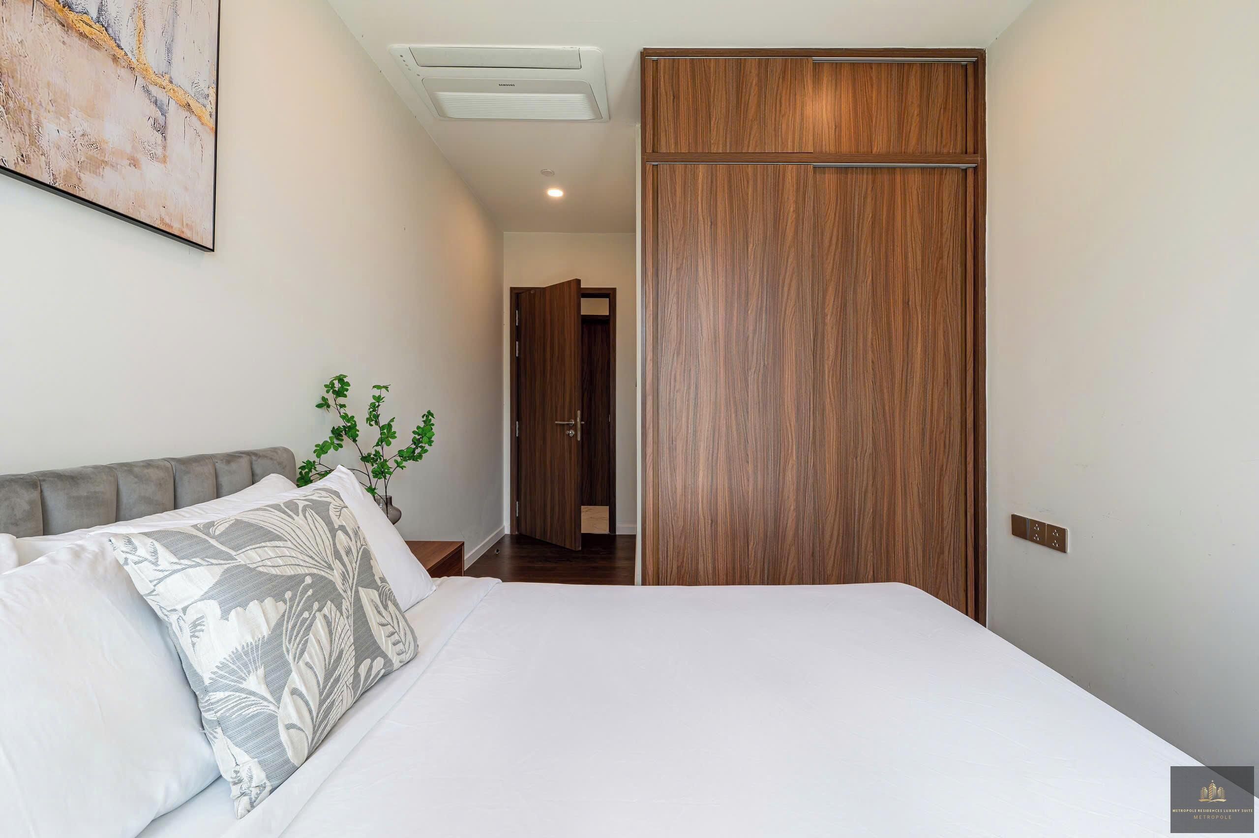 A bedroom with a bed, a wooden wardrobe, a grey headboard, a wall-mounted air conditioner, and a door leading to another room.